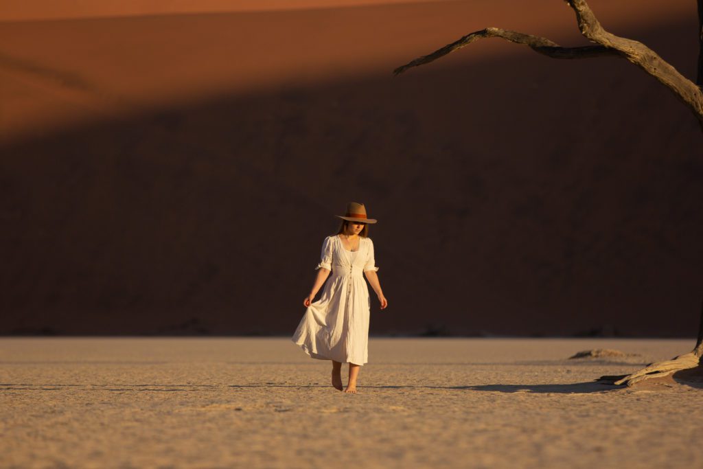 Photographer Emilie Ristevski from @helloemilie walks gracefully across the clay pan of Sossusvlei, with the warm tones of the sand dunes rising majestically behind her.