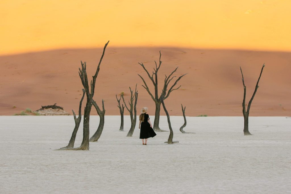 Photographer Emilie Ristevski from @helloemilie in a hat stands amidst the haunting Deadvlei, with its iconic dead trees and the orange dunes of Sossusvlei in the background, embodying the serene solitude of Namibia's desert.
