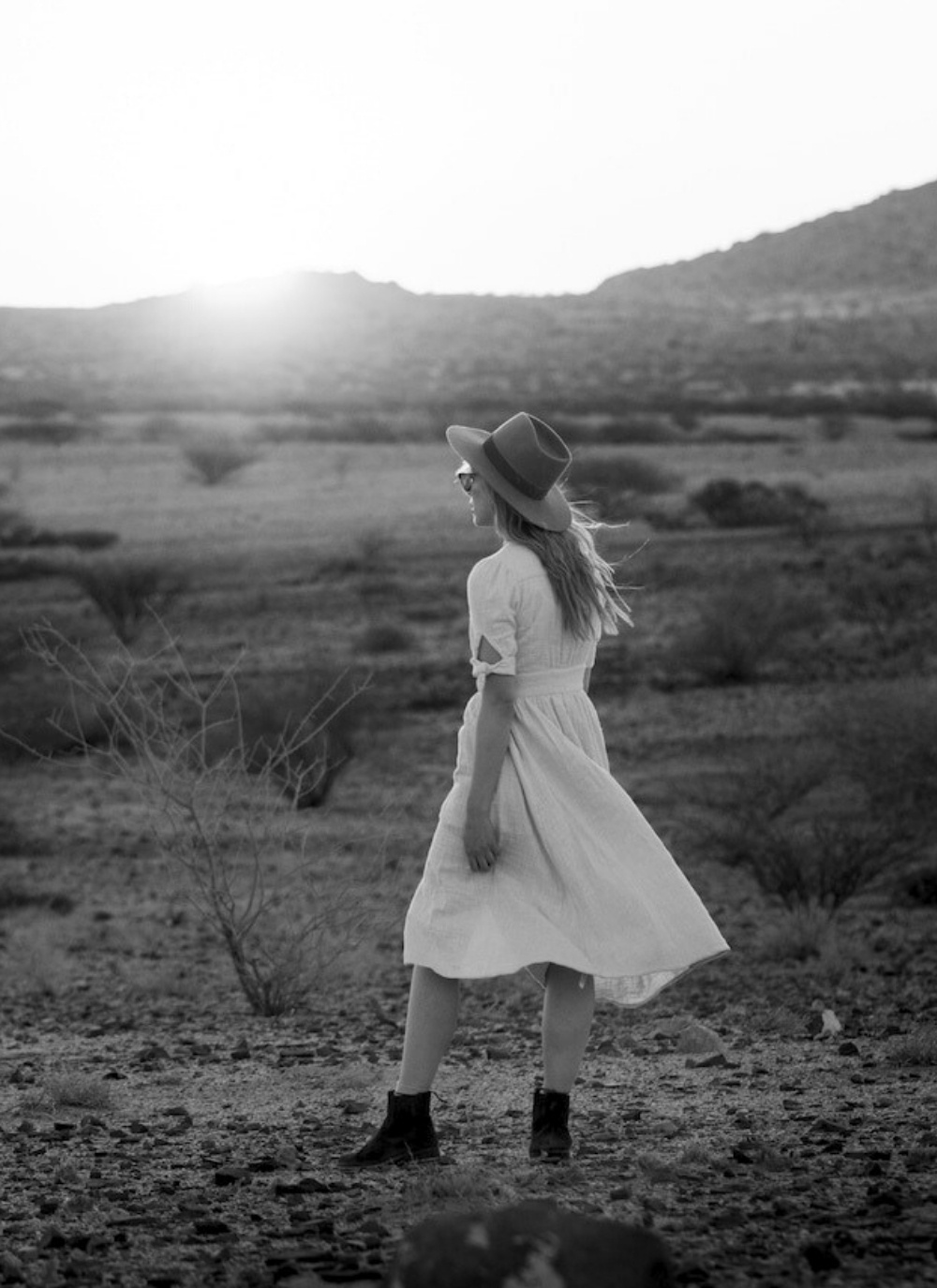Woman in a flowing white dress and hat walking in the golden light of sunset in Etosha National Park, Namibia, with a vast, serene desert landscape in the background.