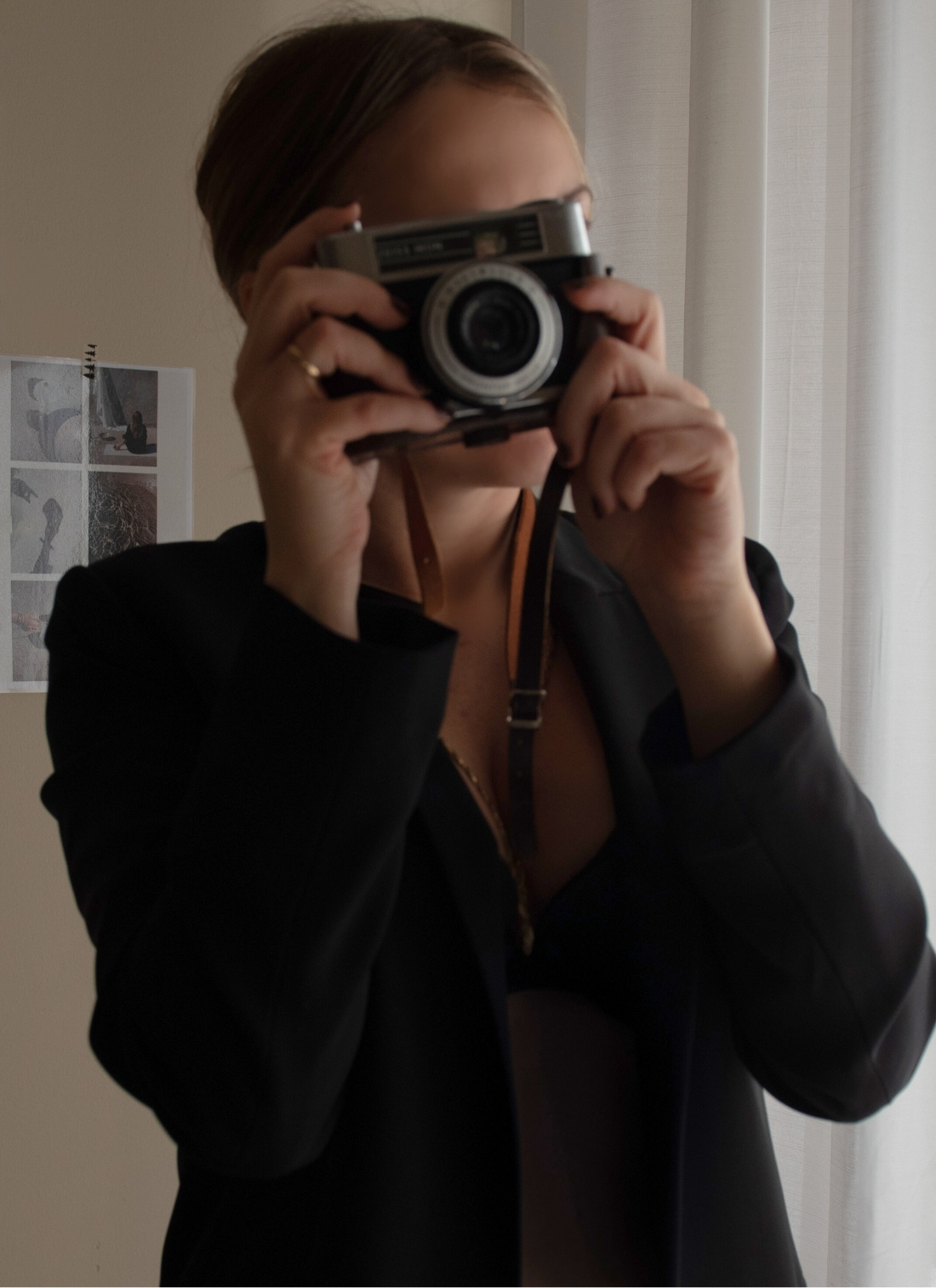 Woman holding a vintage camera in front of her face, standing by soft curtains with a moodboard in the background.