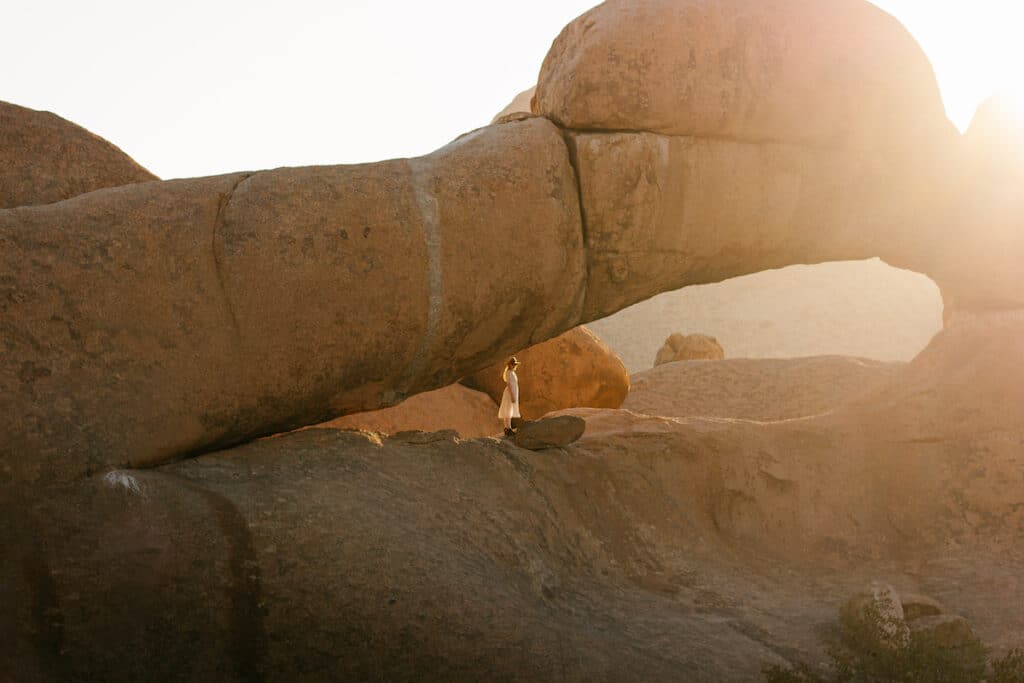 What to visit in Namibia for a week? Spitzkoppe, also known as the 'Matterhorn of Africa,' bathed in the warm glow of the setting sun and Canadese Model Leigh Dickson stands beneath the natural rock arch in Nambia