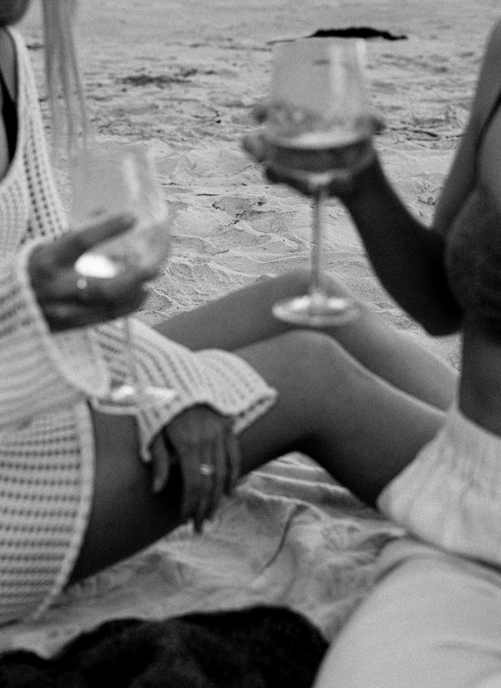 Two women sitting on the beach at sunset, holding wine glasses and sharing a slow, relaxed moment.