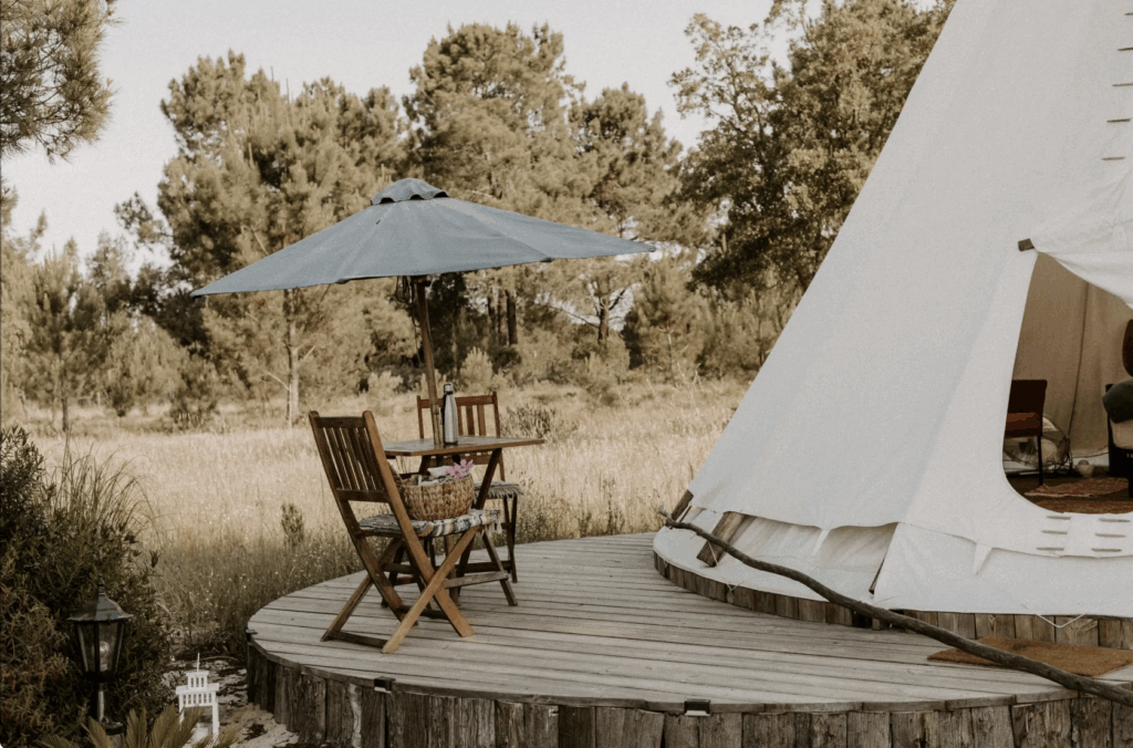 Wooden deck with a teepee tent, shaded table and chairs under a blue umbrella, surrounded by wild grasses and pine trees.