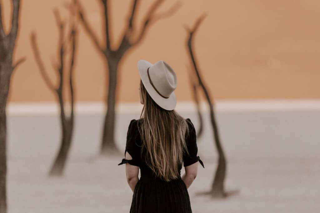 Photographer Emilie Ristevski from @helloemilie stands among the stark and hauntingly beautiful dead acacia trees in the white clay pan of Deadvlei, with the orange dunes of Sossusvlei under a dusky sky in the background.