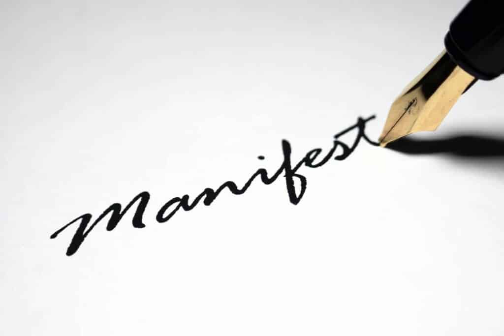 A fountain pen in the process of writing the word 'Manifest' on a white surface, with the 't' at the end unfinished."