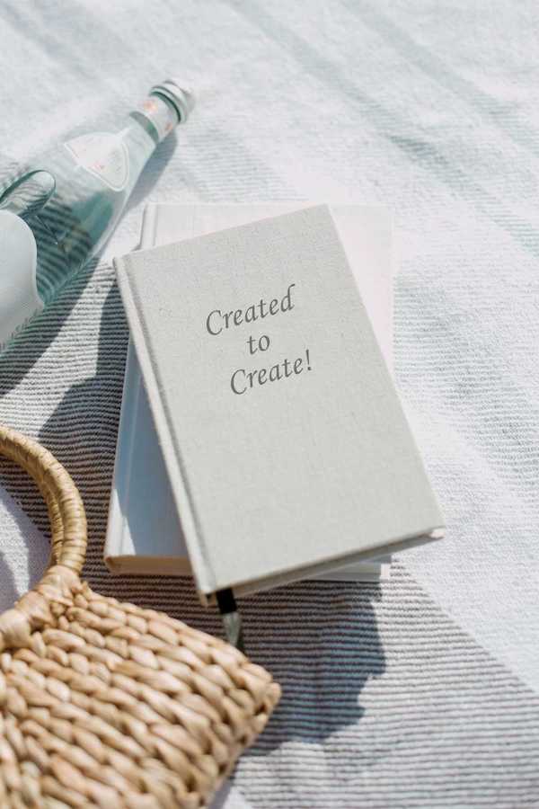 10 best books about manifesting abundance: A journal with 'Created to Create!' on its cover, beside a glass bottle and a woven basket, laid out on a striped cloth under bright sunlight.