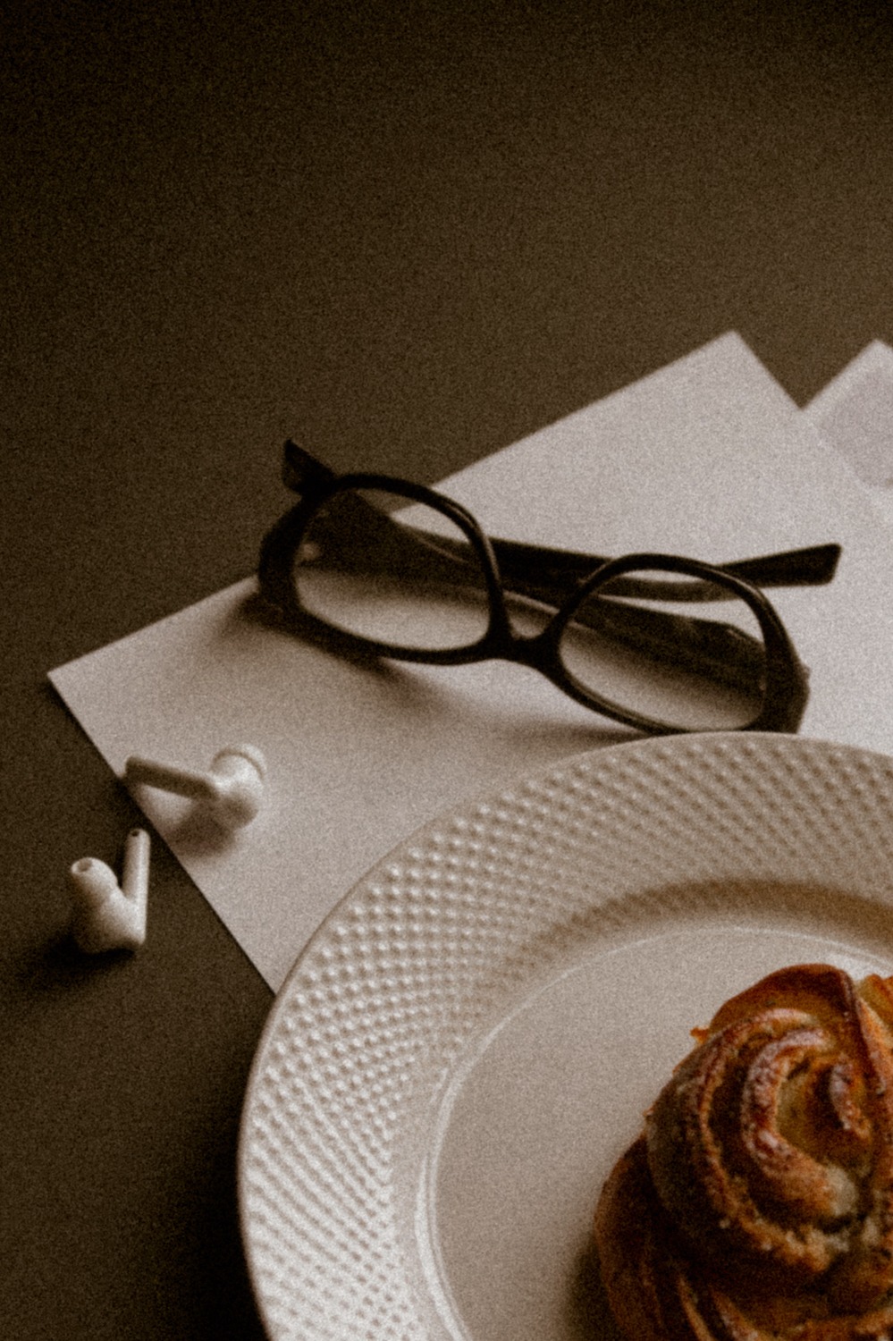 A pair of reading glasses on paper beside a plate with a cinnamon pastry and wireless earphones, in warm vintage light.