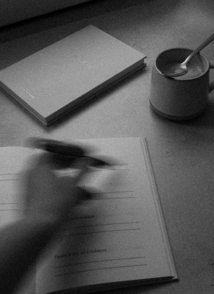 Grainy black-and-white image of a hand journaling next to a cup of coffee and a closed notebook.