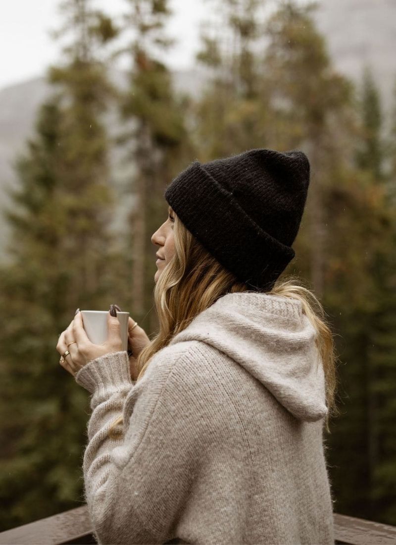 Woman in a wool beanie and cozy sweater enjoying a hot drink on a wooden balcony with forest background, evoking quiet alpine retreat vibes.