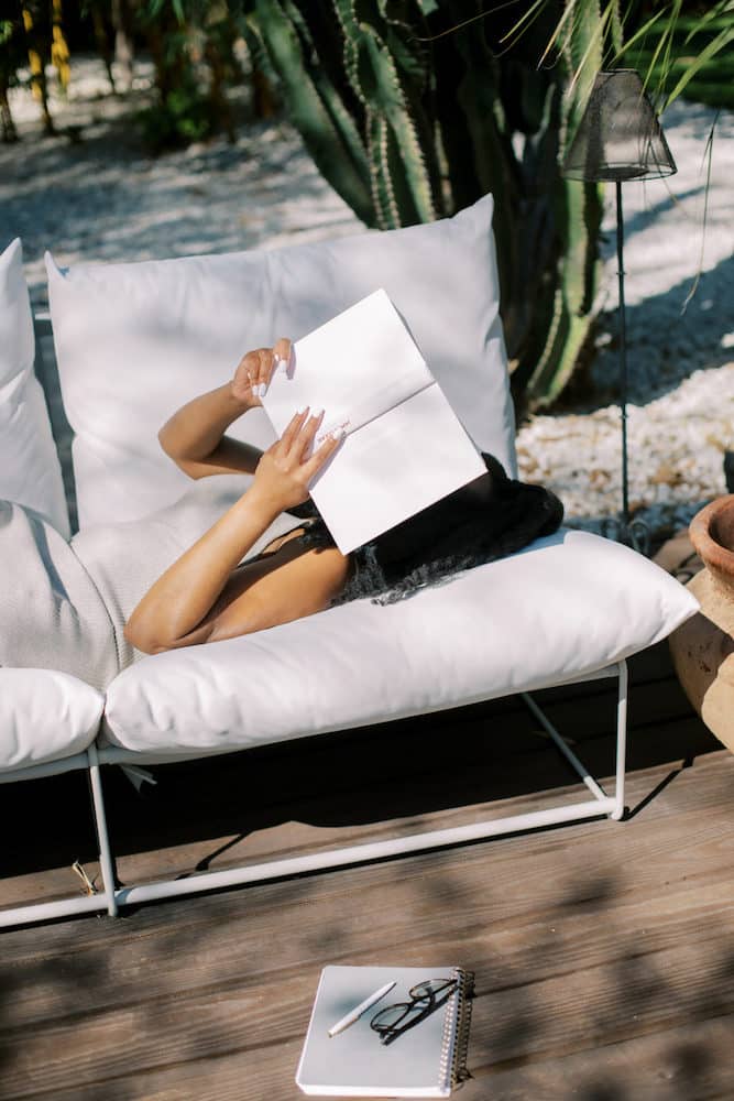 A person leisurely reading books on self-confidence outdoors, reclined on a white cushioned lounge chair, with a notepad and glasses set aside on a wooden deck, embodying a tranquil self-education moment.
