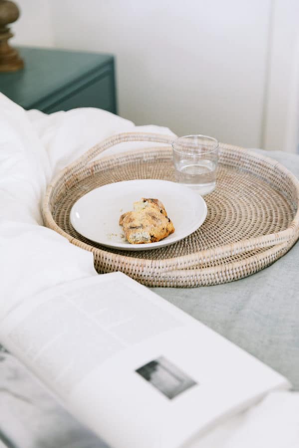 Breakfast in bed featuring a half-eaten scone on a plate, a glass of water, and an open book on a woven tray, suggesting a relaxed morning reading of the best books on confidence.
