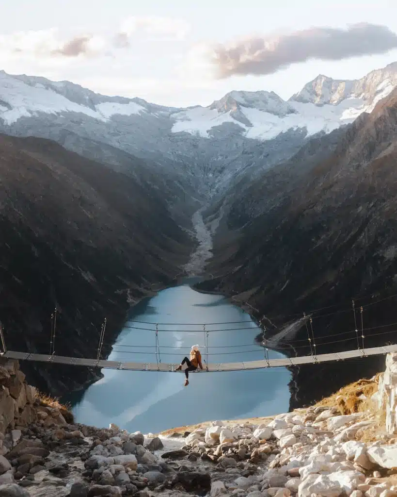 The Blisshunter sits midway on a suspension bridge over a glacial lake on the approach to Olpererhütte Austria, surrounded by the dramatic Zillertal Alps.