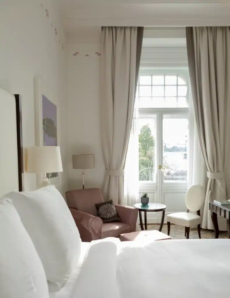 A bright and airy hotel room in the Four Seasons Budapest with elegant décor, featuring a plush armchair, a large bed, and tall draped windows overlooking the city.