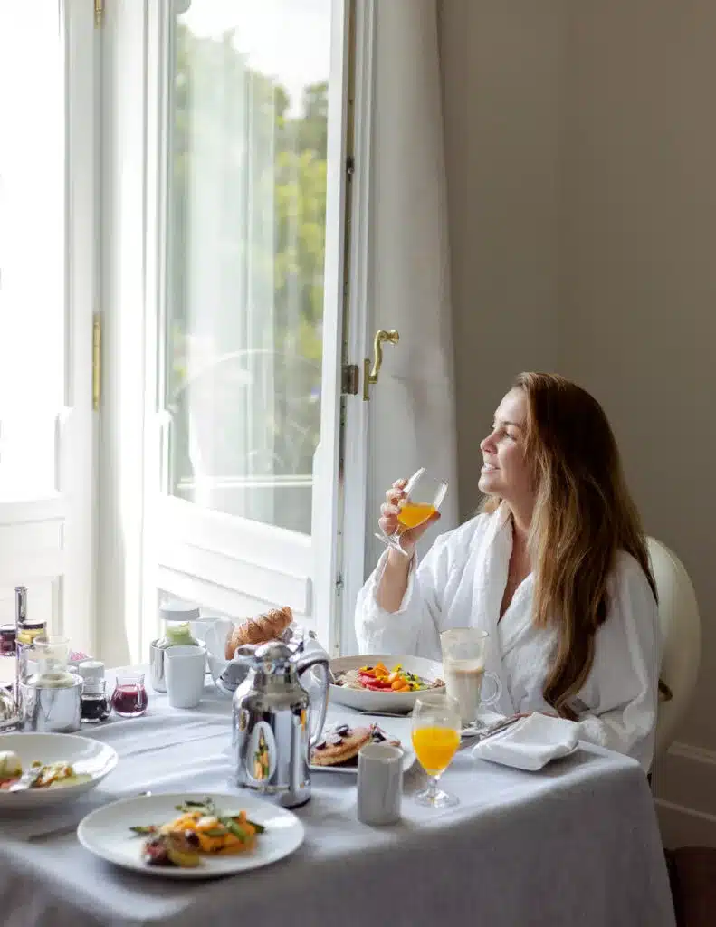 The Blisshunter in a white robe enjoys a luxurious breakfast by the window in a sunlit hotel room at the Four Seasons Budapest Hotel, with a view of lush greenery outside.