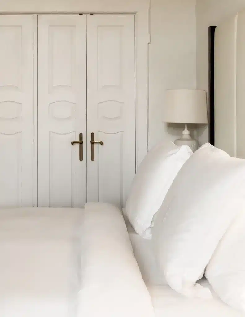 A close-up of a luxurious hotel bed at Four Seasons Budapest with fluffy white pillows, a crisp white duvet, and an elegant bedside lamp, with classic white double doors in the background.