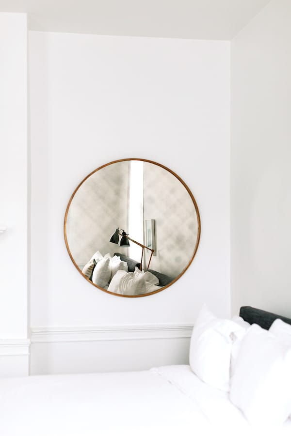 A modern bedroom reflected in a large round mirror with a thin wooden frame, capturing the crisp white bedding and plush pillows.