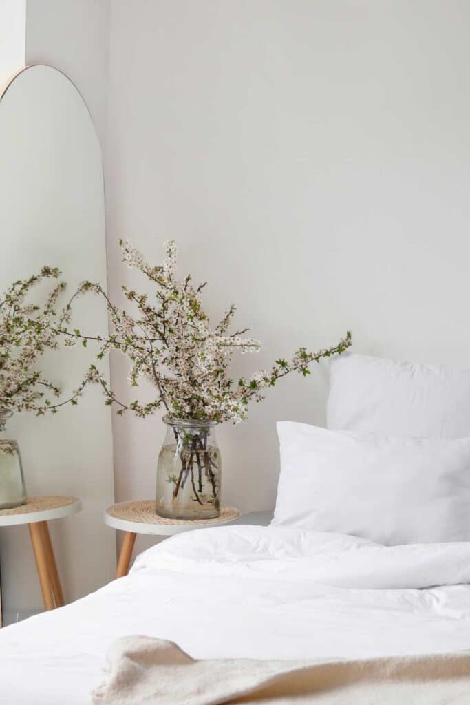 A serene bedside setting featuring a glass vase with delicate white blossoms on a wooden side table next to a bed with white linen.