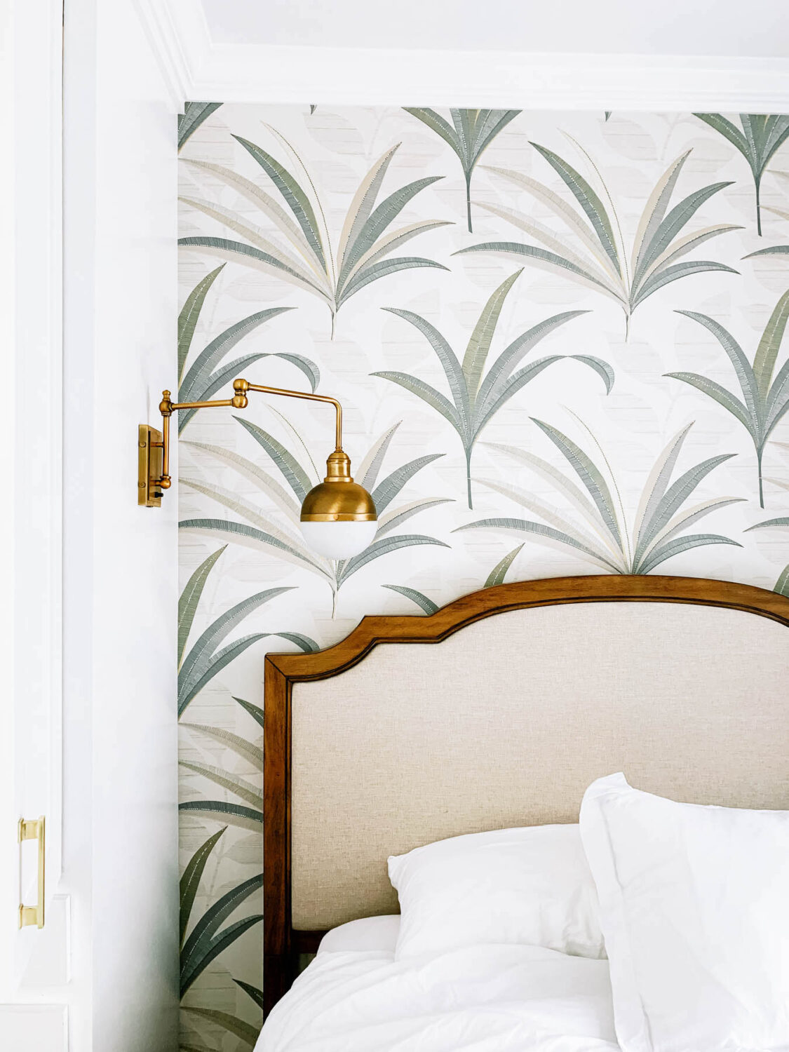 Part of a luxurious bedroom showing a portion of an upholstered bed with a wooden frame, against a backdrop of tropical leaf-patterned wallpaper, complemented by a brass wall-mounted reading light - 10 Easy Styling tips for a Luxury Bedroom makeover -