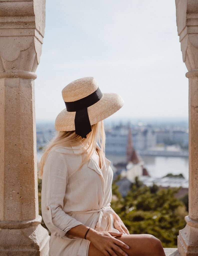Woman in a straw hat overlooking the Budapest cityscape from an arched balcony of Fisherman's Bastion.