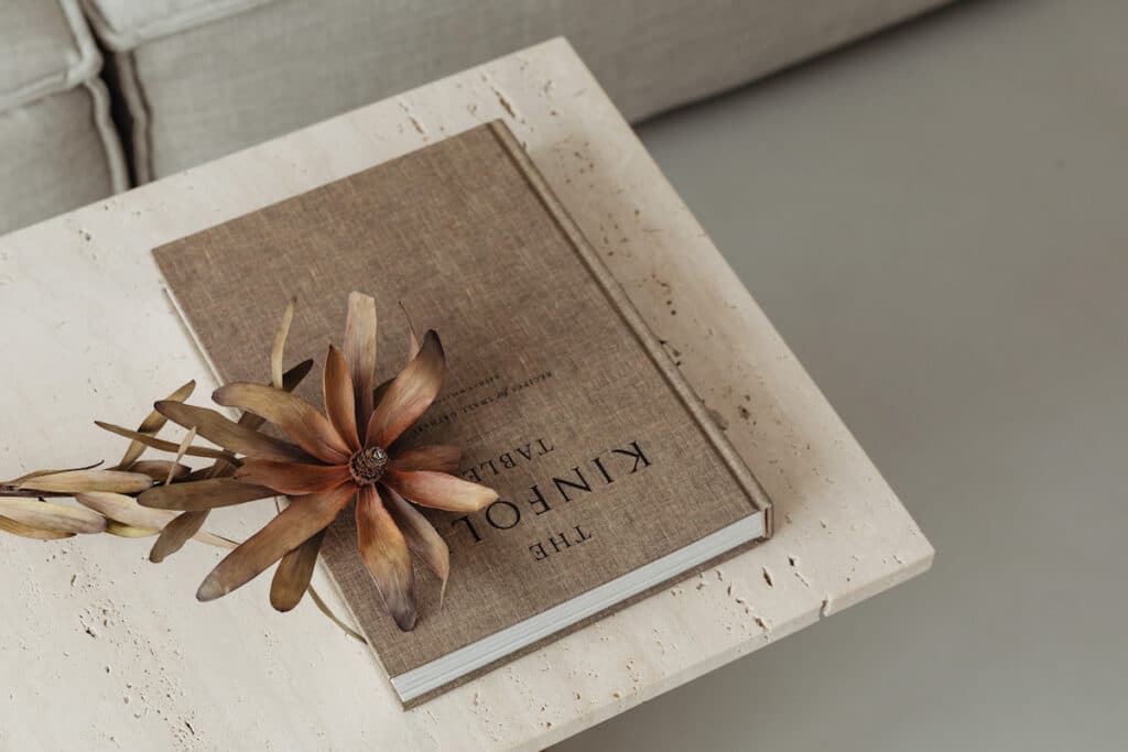 Kinfolk Luxury coffee table books with a linen cover rests on a white stone table, accompanied by a dried lotus flower, embodying sophisticated home decor.