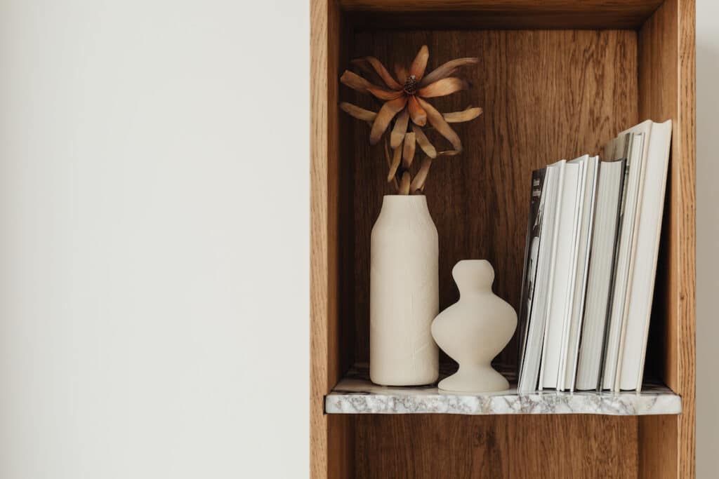  beige textured vase with a dried flower arrangement stands on a marble shelf, complemented by a sculptural piece, within a wooden alcove, luxury coffee table books epitomizing luxury home decor.