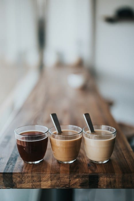 Three different healthy coffee alternative beverages displayed in clear glasses on a long wooden bar table.