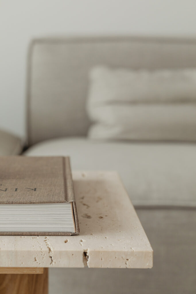 A close-up of textured luxury coffee table books on a stone table, with a soft-focus background of a cozy, modern grey sofa, illustrating a tranquil reading nook