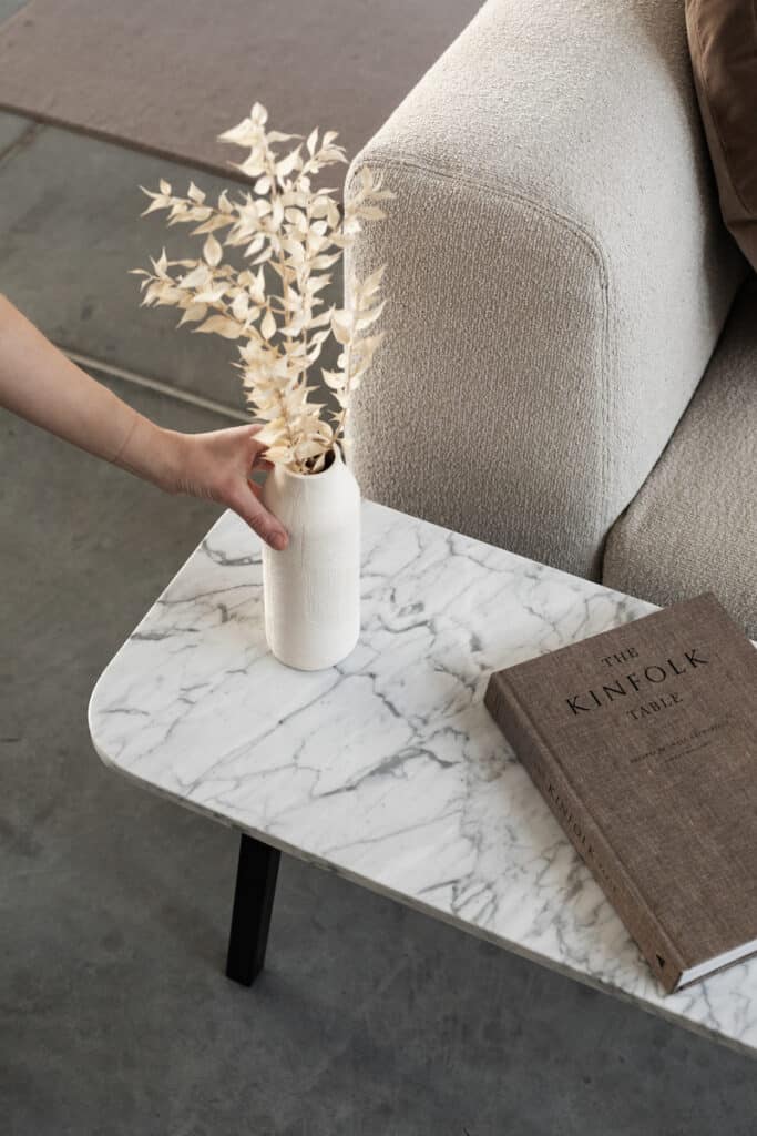 A modern marble coffee table featuring one of the luxury books titled "The Kinfolk Table" alongside a white vase with dried foliage, held by a hand.