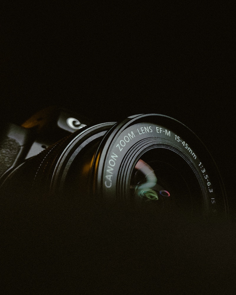 A close-up of a EF Canon zoom lens in a moody, dark setting with a soft light highlighting the details and text on the lens barrel.