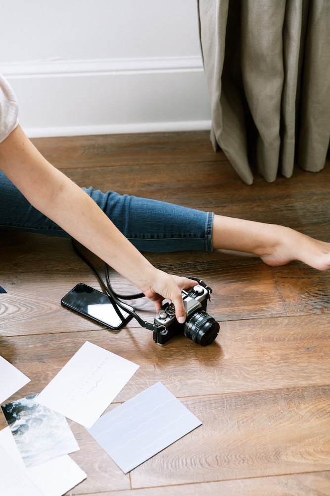 A photographer's casual workspace with a DSLR camera in hand, surrounded by scattered travel photos on a wooden floor, symbolizing the selection process for the best lens for travel photography.