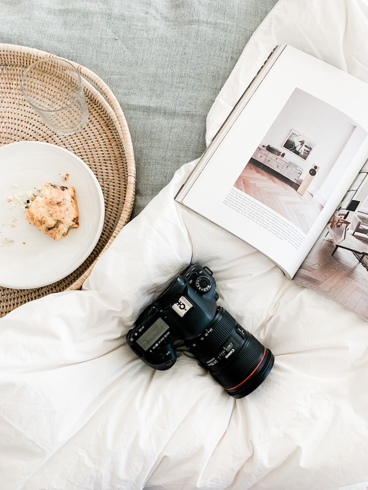 An open photography book lies next to a professional camera with a red-ringed lens and a half-eaten scone on a plate, capturing the essence of a photographer's relaxed morning choosing the best lens for travel photography.