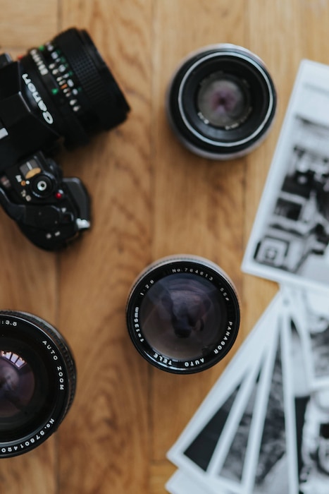 Best lens for travel photography: An assortment of camera lenses and a DSLR camera surrounded by black and white travel photographs on a wooden surface.
