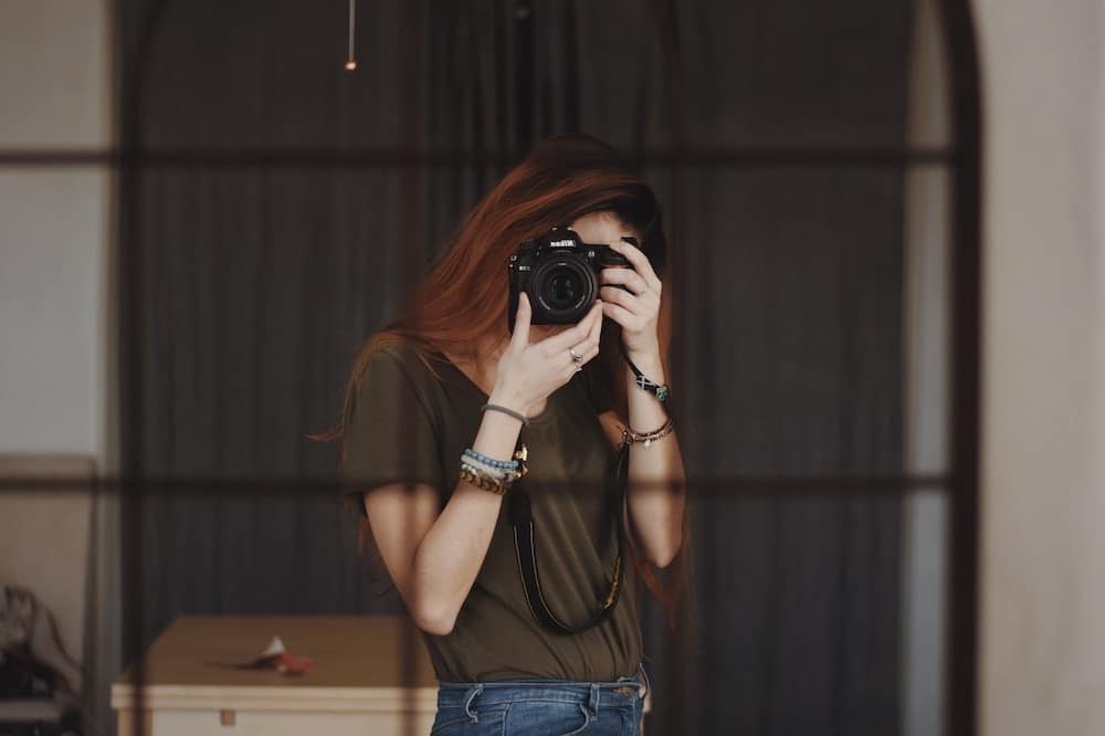 A photographer with auburn hair, in a casual olive tee and jeans, is focused through Best Lens for Travel Photography of her DSLR camera, capturing the essence of the moment, set against the backdrop of a softly lit room with elegant drapery.