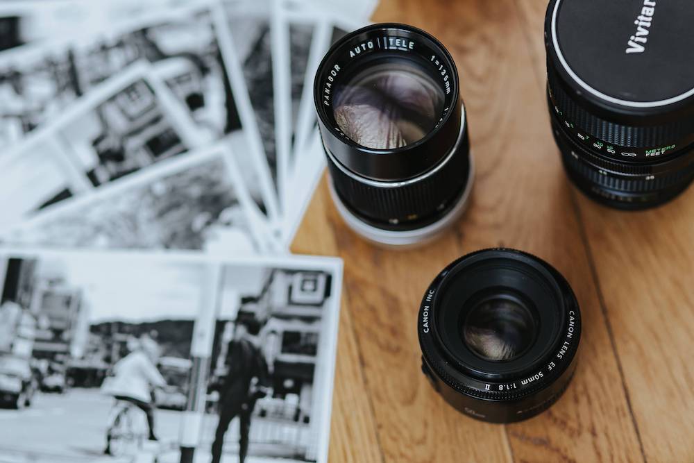 A close-up of professional camera lenses and black and white street photography prints strewn across a wooden surface, depicting the essence of urban travel photography - The Art of Choice: Finding the Best Lens for Urban Travel Photography