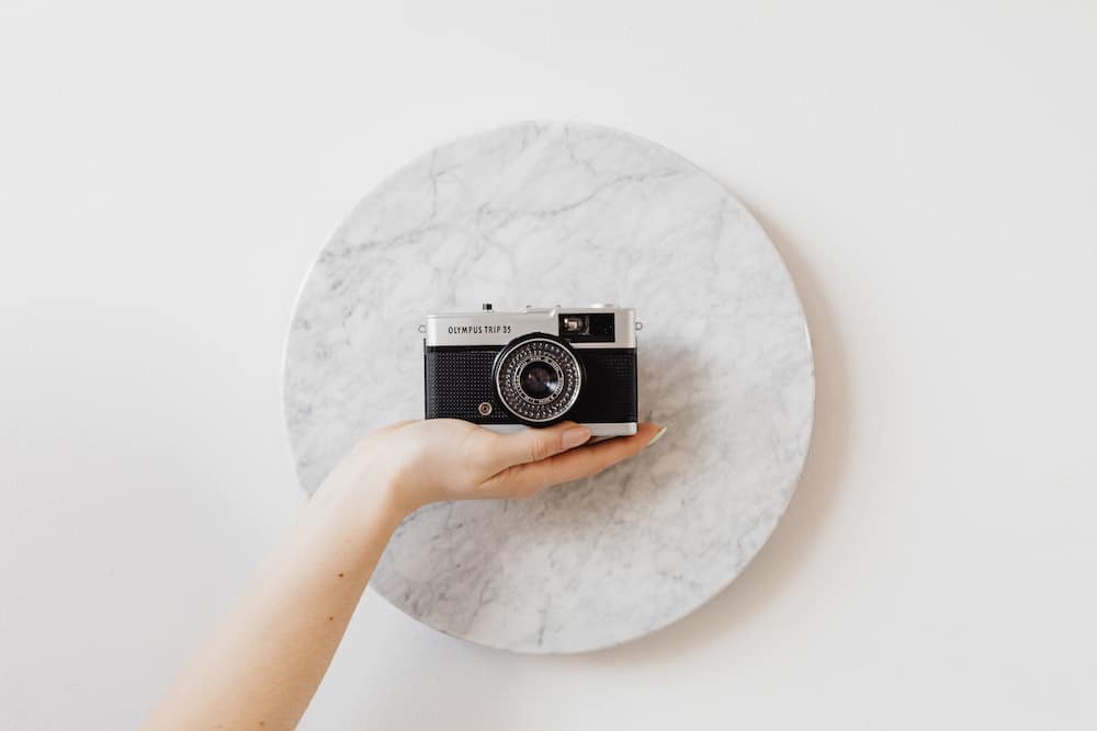 A hand gracefully holds a vintage Olympus Trip 35 camera against the backdrop of a circular marble slab, contrasting the timeless elegance of classic photography gear with a modern minimalist setting. - Capturing Timelessness: Choosing the Best Lens for Travel Photography