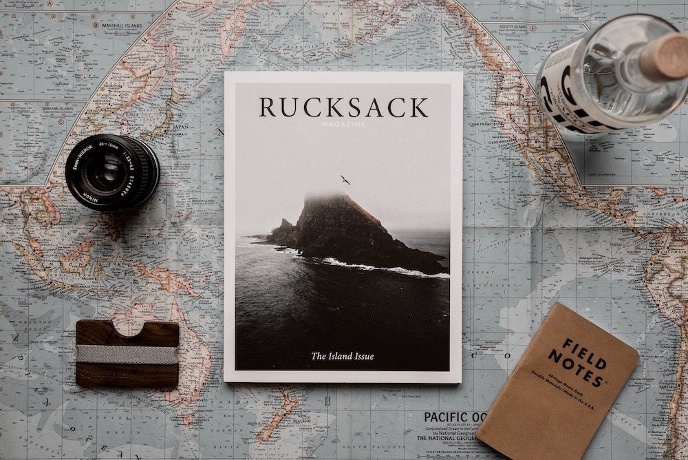 A traveler's flat lay composition with a camera lens atop a detailed world map, beside 'RUCKSACK Magazine' featuring a misty seascape on its cover, and a 'Field Notes' notebook, evoking the spirit of adventure and photography.