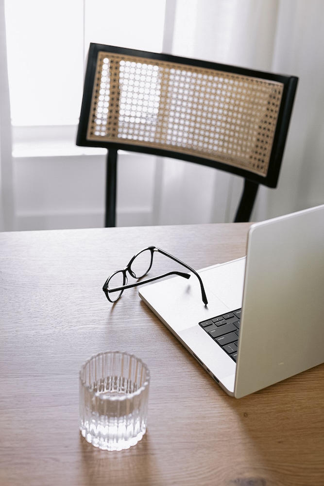 A minimalist workspace featuring an open laptop on a wooden desk, with a pair of black-rimmed glasses set aside and a clear glass of water, all illuminated by natural light from a window beside a stylish chair with a woven back. Perfect place for reading Confidence Books.