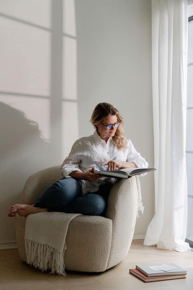 A contemplative woman sits in a cozy, oversized chair, deeply engrossed in Confidence Books. She's dressed in a casual white blouse and denim jeans, with sunlight casting geometric shadows through a nearby window, enhancing the peaceful ambiance of a quiet reading corner.