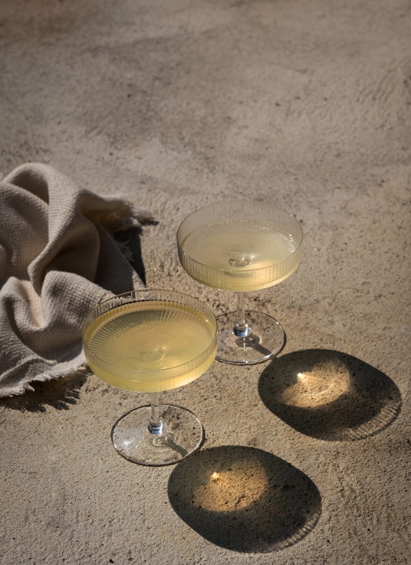 Two elegant cocktail glasses filled with a light drink casting delicate shadows on a sunlit sandy surface, next to a soft beige cloth.