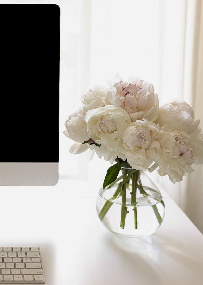 A vase of white peonies on a desk next to a computer and keyboard, symbolizing serenity and inspiration for reading books about feminine energy.