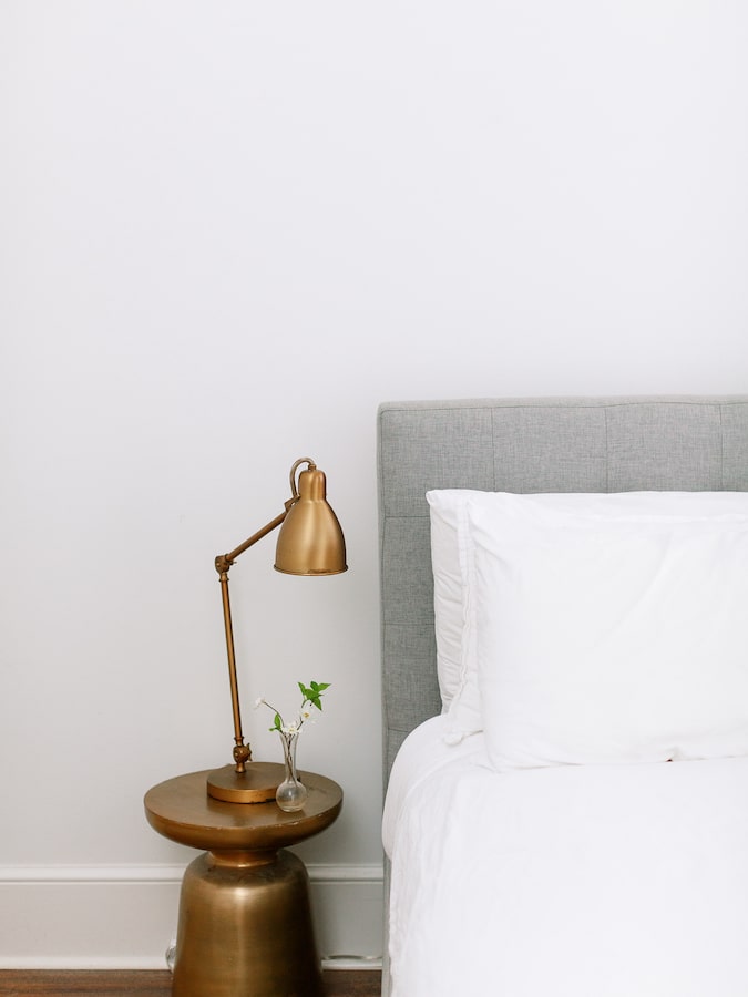 A minimalist bedroom scene with a grey upholstered headboard, crisp white bedding, a brass table lamp, and a round bedside table in a matching brass finish with a delicate vase holding a sprig.
