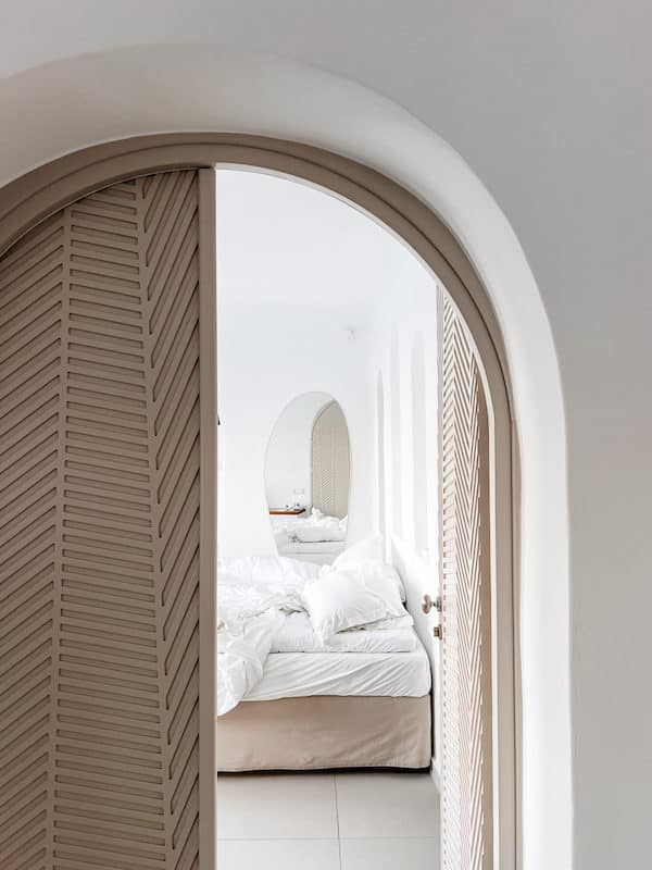 View through an arched doorway revealing a luxury bedroom makeover with crisp white bedding and louvered closet doors, creating a serene and stylish sleeping space.