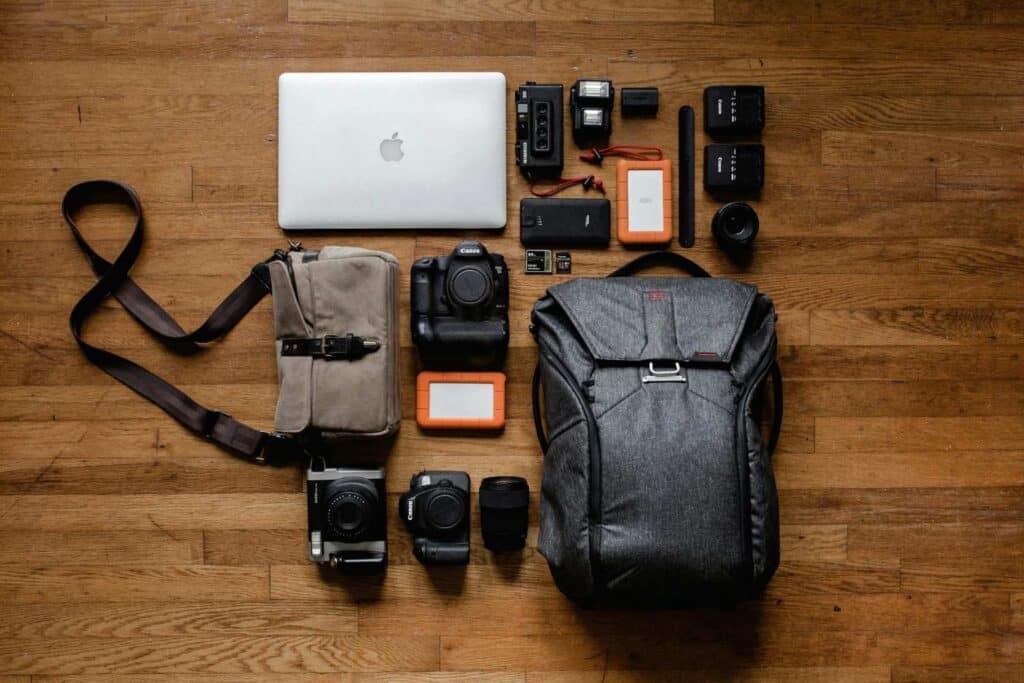 A meticulously organized array of travel photography gear, including cameras, lenses, and a laptop, peak design - one of the best camera bags for travel laid out beside a durable camera bag on a wooden floor.