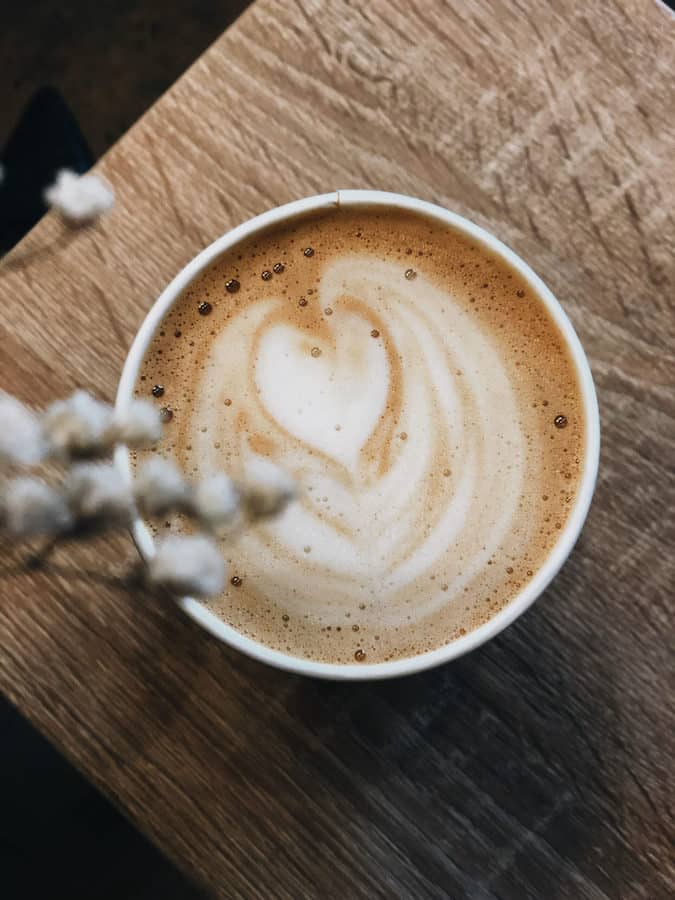 Top-down view of a latte with heart-shaped latte art on a wooden surface, accompanied by a sprig of dried flowers.