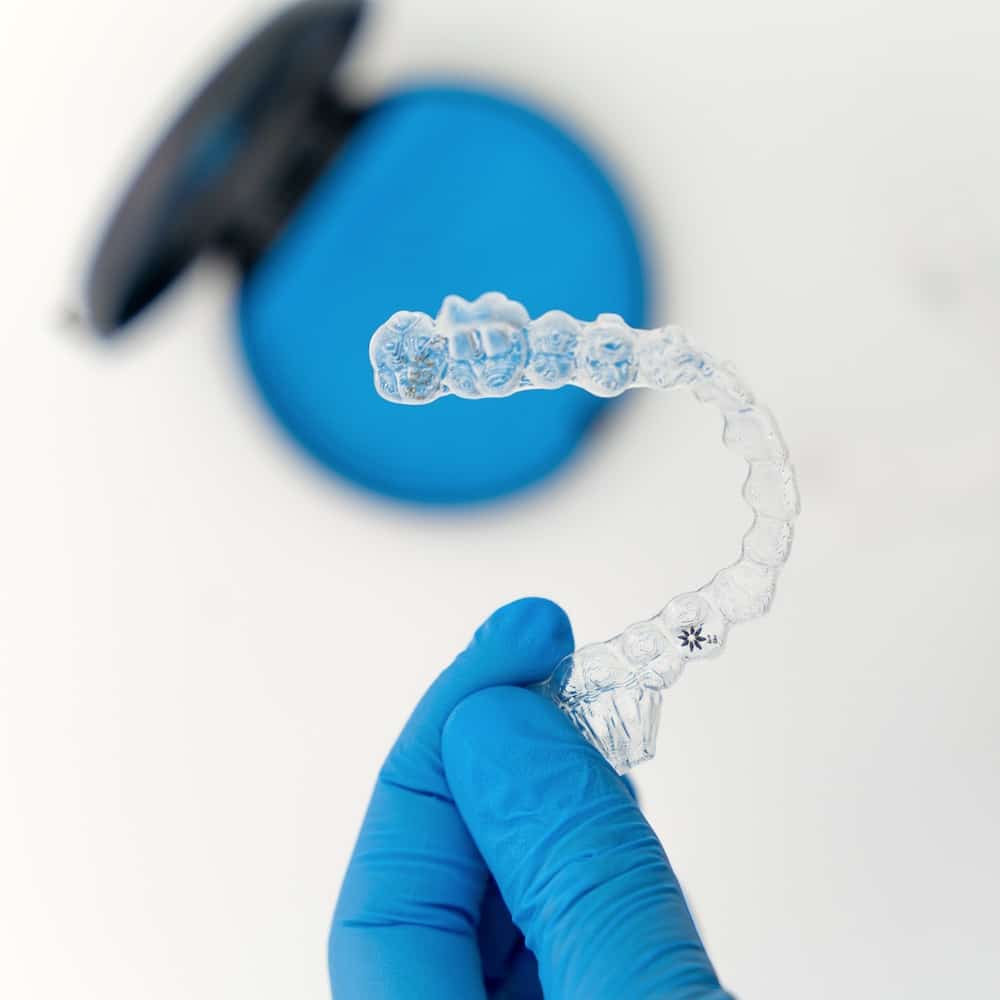A gloved hand carefully holds a transparent Invisalign aligner above a blue case, highlighting the aligner's intricate details.