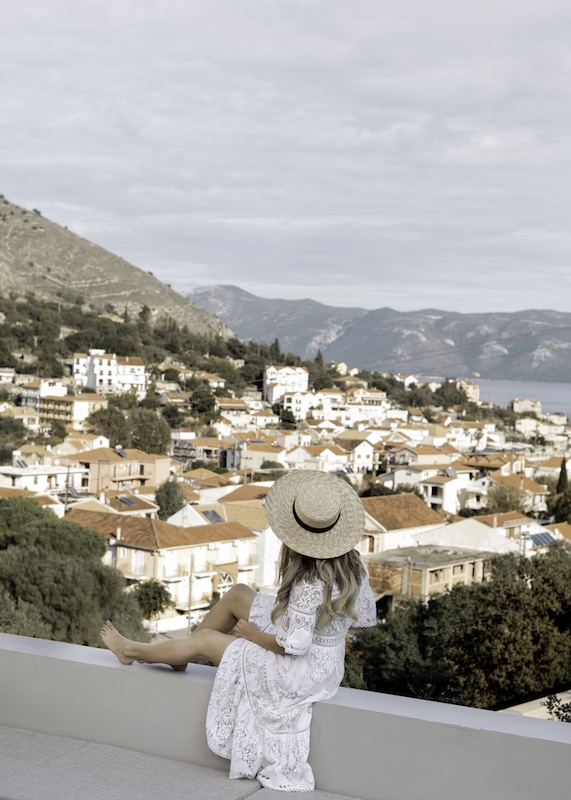 A woman in a flowing white dress and straw hat sits overlooking the terracotta rooftops of a Cephalonian town, with the Ionian Sea in the distance.