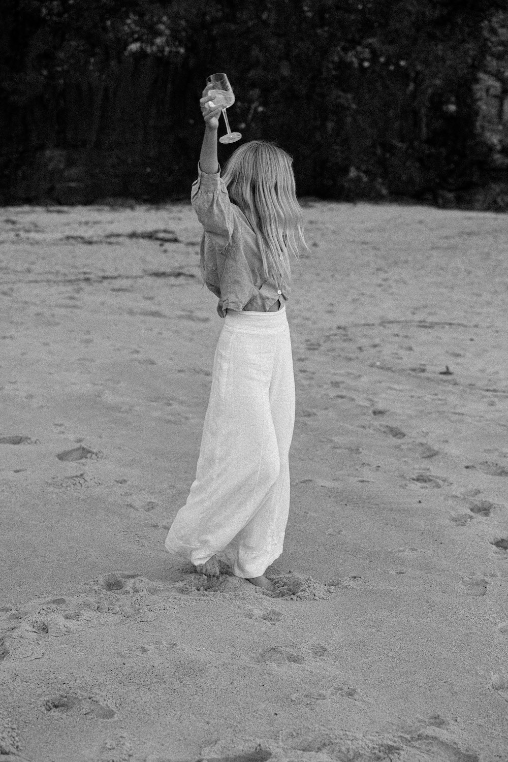 Barefoot woman in wide-leg linen pants holding wine glass on beach — carefree, confident moment in black and white