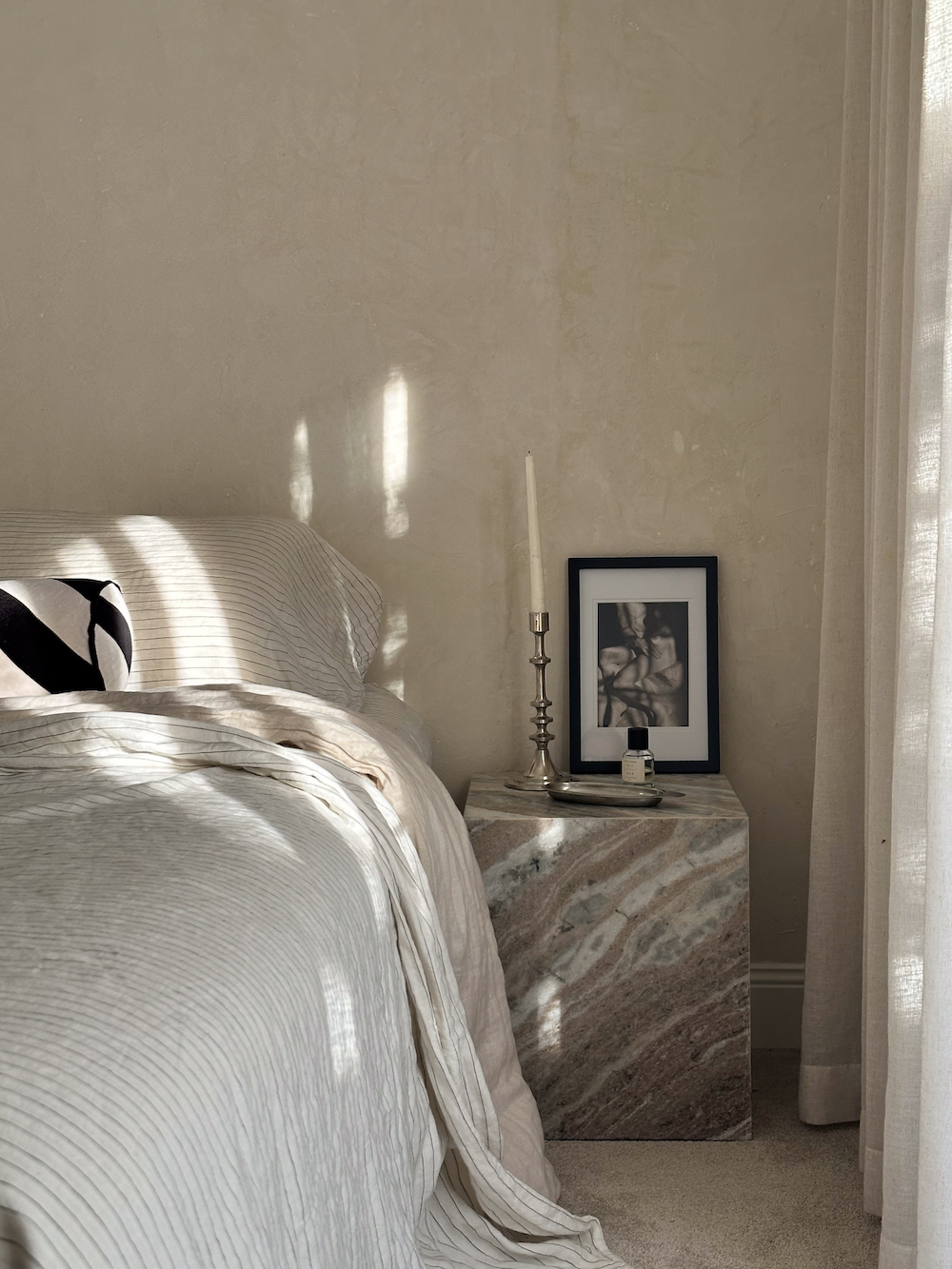 A serene bedroom corner features a neatly made bed with light textiles and a stylish nightstand adorned with a candle, a framed artwork, and delicate shadows from sunlight.