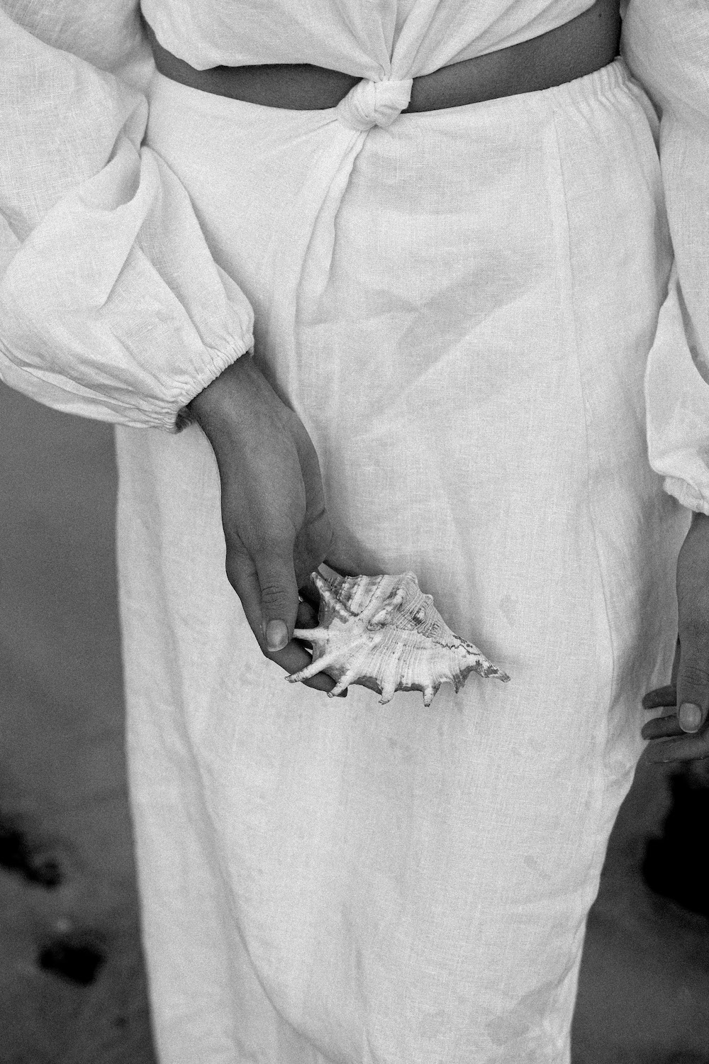 A person dressed in a flowing white ensemble delicately cradles a beautiful conch shell while standing on sandy shores as the soft evening light casts gentle shadows.