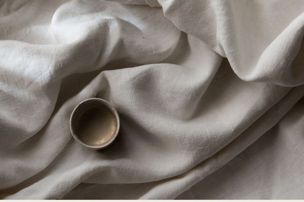 Ceramic cup resting on softly crumpled natural linen bedding, evoking calm, warmth, and slow living.